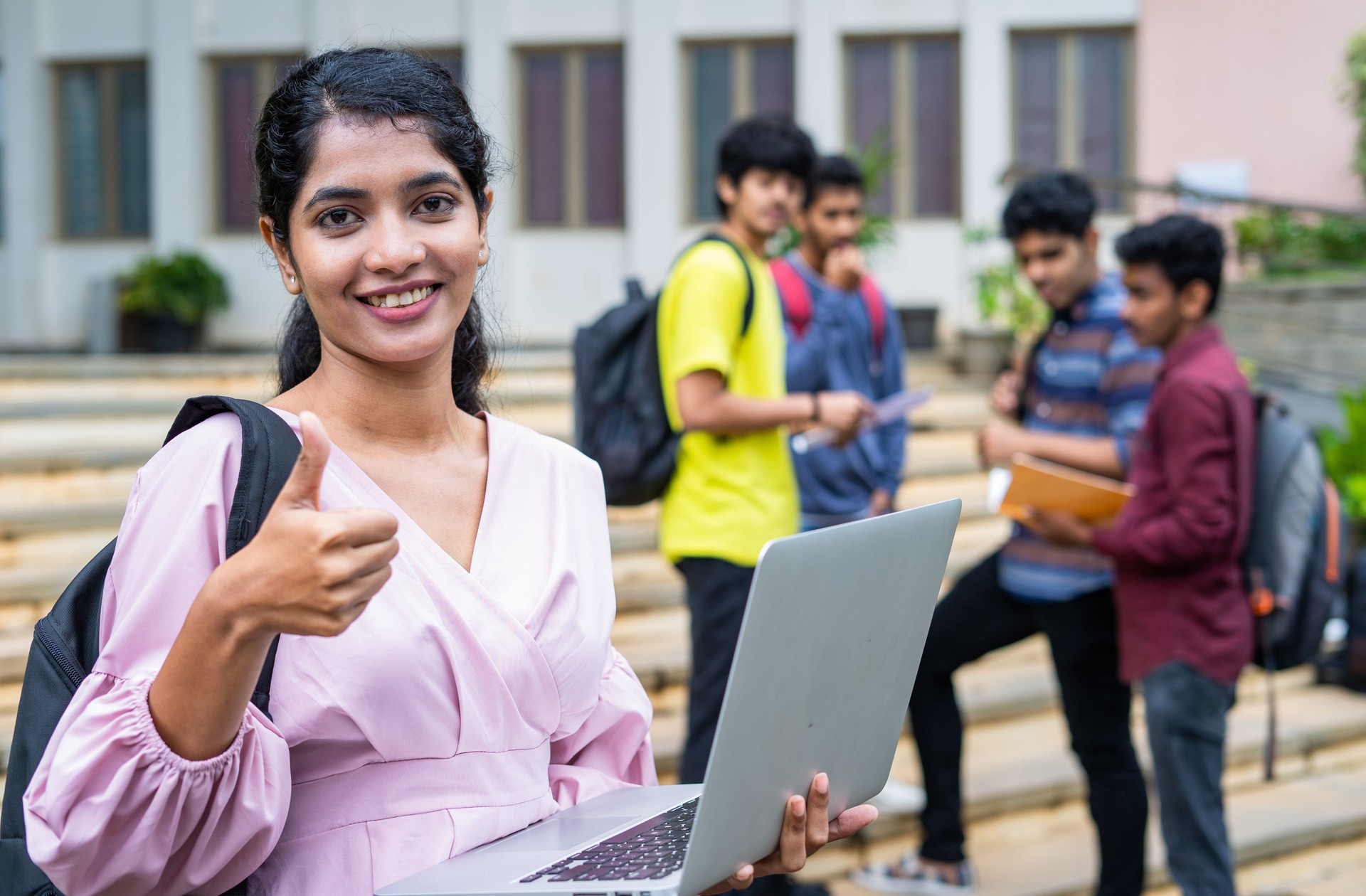 confidence indian girl student showing thumbs up by looking at camera at college campus while using laptop -concept of education promotion, successful and advertisement